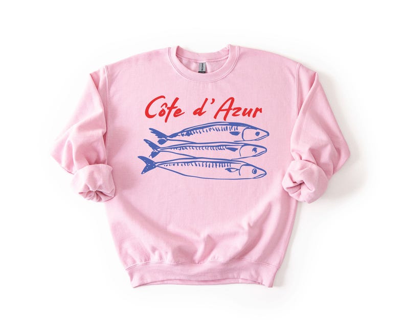 K&ouml;nnte beinhalten: Ein hellrosa Sweatshirt mit hochgekrempelten &Auml;rmeln. Auf der Vorderseite steht der Text "C&ocirc;te d'Azur" in Rot &uuml;ber drei blauen Fischillustrationen. Es ist ein l&auml;ssiges Kleidungsst&uuml;ck.