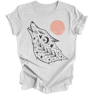 May include: A white t-shirt featuring a black and white illustration of a wolf howling at a pink sun. The wolf is adorned with stars, leaves, and a crescent moon.