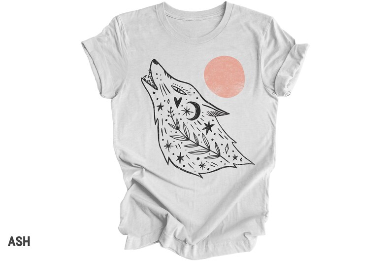 May include: A white t-shirt featuring a black and white illustration of a wolf howling at a pink sun. The wolf is adorned with stars, leaves, and a crescent moon.
