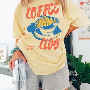 Puede incluir: Camiseta amarilla con un gr&aacute;fico de una taza de caf&eacute; con un croissant en la parte superior. El texto de la camiseta dice "Coffee Club" y "Everyday All Day".