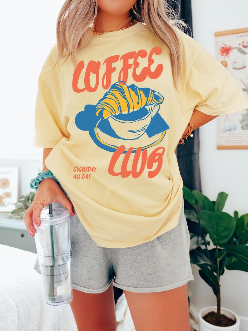 Puede incluir: Camiseta amarilla con un gr&aacute;fico de una taza de caf&eacute; con un croissant en la parte superior. El texto de la camiseta dice "Coffee Club" y "Everyday All Day".