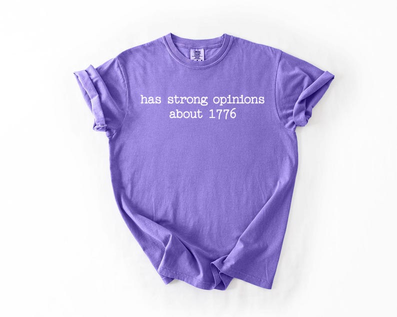 Puede incluir: Una camiseta lavanda con el texto blanco "has strong opinions about 1776". La camiseta tiene las mangas enrolladas y est&aacute; hecha de un material suave. La camiseta se muestra sobre un fondo blanco.