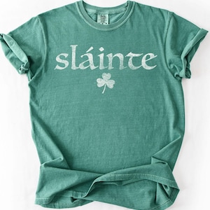 May include: A teal green t-shirt with the word "sl&aacute;inte" in white, stylized lettering and a white shamrock graphic. The shirt has a crew neck and short sleeves. The fabric appears soft and slightly faded.
