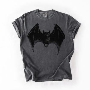 May include: Dark gray t-shirt featuring a black bat graphic with outstretched wings. The bat is centered on the shirt, with detailed wing and body features. The shirt has a vintage, worn look.