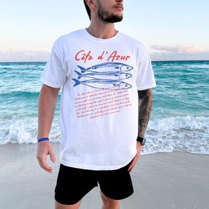 May include: White t-shirt with the text "C&ocirc;te d'Azur" in red and blue fish illustrations. The shirt also features a paragraph of text in red. The person is wearing black shorts and a blue bracelet, standing on a beach with the ocean in the background.