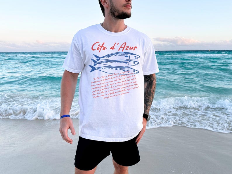 May include: White t-shirt with the text "C&ocirc;te d'Azur" in red and blue fish illustrations. The shirt also features a paragraph of text in red. The person is wearing black shorts and a blue bracelet, standing on a beach with the ocean in the background.