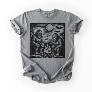 May include: Gray t-shirt featuring a black and white graphic of two Bigfoot creatures roasting marshmallows over a campfire under a full moon. The design includes bats flying in the sky. The shirt has a classic crew neck and short sleeves.