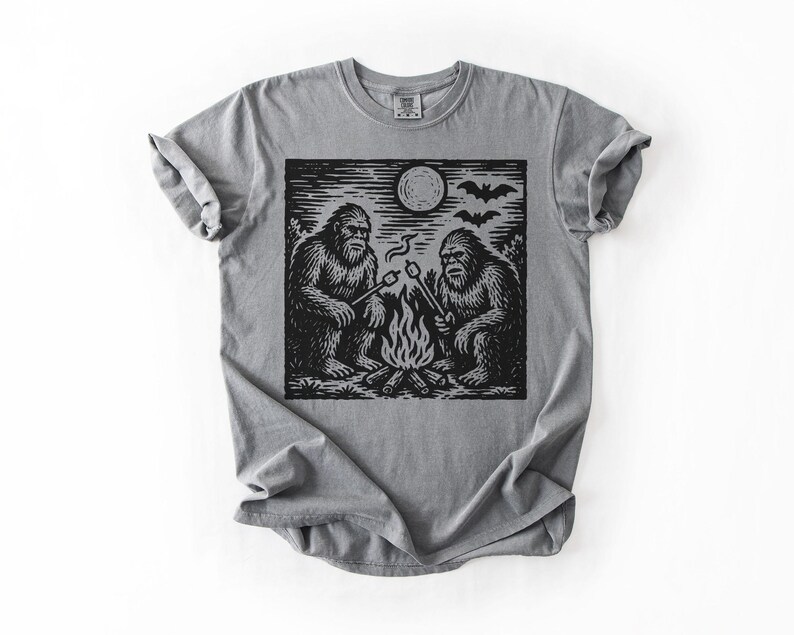 May include: Gray t-shirt featuring a black and white graphic of two Bigfoot creatures roasting marshmallows over a campfire under a full moon. The design includes bats flying in the sky. The shirt has a classic crew neck and short sleeves.