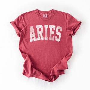 May include: A red t-shirt with the word "ARIES" printed in white, block letters. The shirt has rolled-up sleeves and a crew neck. The fabric has a slightly faded appearance.