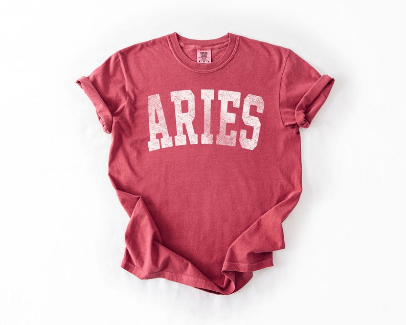 May include: A red t-shirt with the word "ARIES" printed in white, block letters. The shirt has rolled-up sleeves and a crew neck. The fabric has a slightly faded appearance.