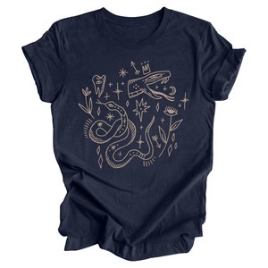 Puede incluir: Una camiseta azul marino con un dise&ntilde;o gr&aacute;fico beige que presenta una serpiente, un diente, una corona, estrellas y un ojo.
