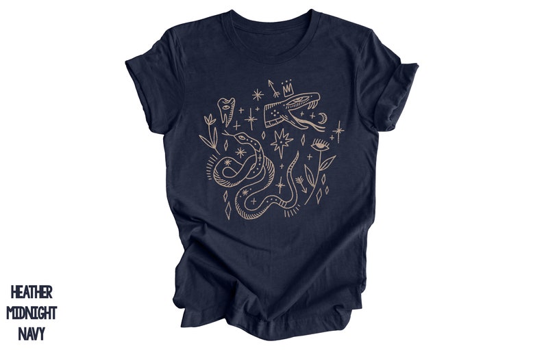 Puede incluir: Una camiseta azul marino con un dise&ntilde;o gr&aacute;fico beige que presenta una serpiente, un diente, una corona, estrellas y un ojo.