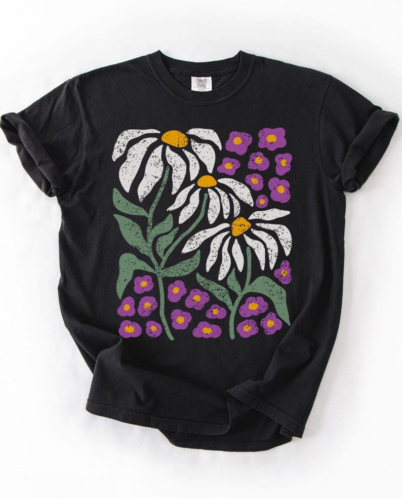 May include: Black t-shirt featuring a floral design. The graphic includes three large white daisies with yellow centers, green stems and leaves, and small purple flowers. The shirt has a classic crew neck and short sleeves.