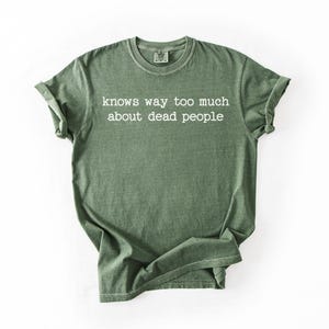 Puede incluir: Camiseta verde oliva con el texto blanco "knows way too much about dead people". La camiseta tiene cuello redondo y mangas enrolladas. La tela parece suave y ligeramente descolorida.