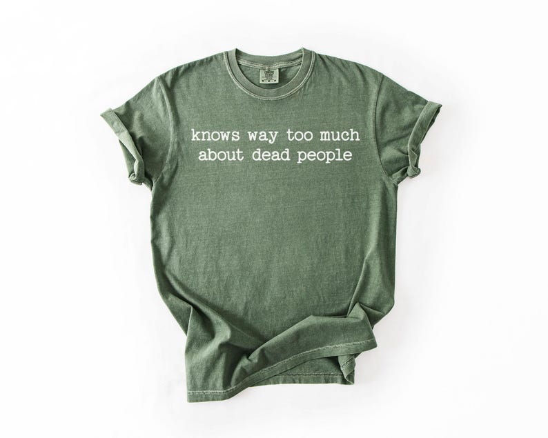 Puede incluir: Camiseta verde oliva con el texto blanco "knows way too much about dead people". La camiseta tiene cuello redondo y mangas enrolladas. La tela parece suave y ligeramente descolorida.