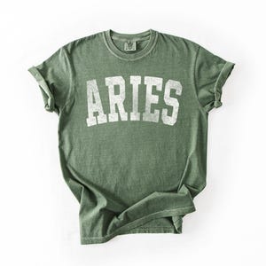 May include: An olive green t-shirt with the word "ARIES" printed in white, centered on the front. The shirt has a vintage, slightly faded look, with rolled-up sleeves. The fabric appears soft and comfortable.