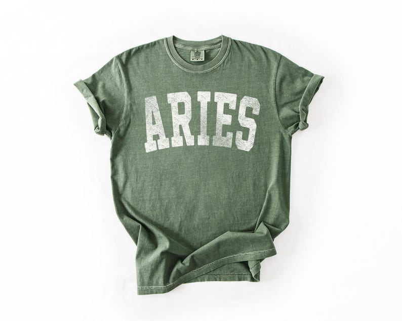 May include: An olive green t-shirt with the word "ARIES" printed in white, centered on the front. The shirt has a vintage, slightly faded look, with rolled-up sleeves. The fabric appears soft and comfortable.