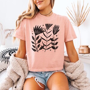 May include: A pale pink t-shirt with a black floral design. The graphic features stylized flowers, leaves, and stars. The short-sleeved shirt is made of soft material. The model is wearing denim shorts and a cream-coloured sweater.