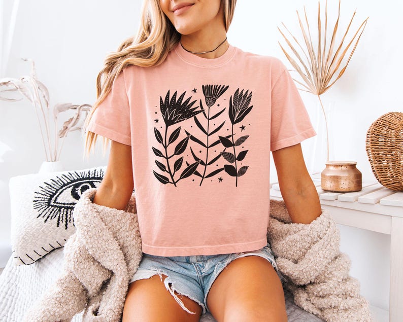 May include: A pale pink t-shirt with a black floral design. The graphic features stylized flowers, leaves, and stars. The short-sleeved shirt is made of soft material. The model is wearing denim shorts and a cream-coloured sweater.