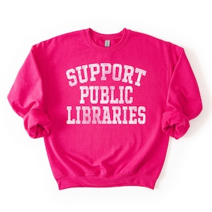 May include: A bright pink sweatshirt with the words "SUPPORT PUBLIC LIBRARIES" printed in white, arched text. The sweatshirt has long sleeves and a crew neck. The text has a distressed, vintage look.