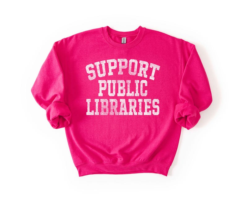 May include: A bright pink sweatshirt with the words "SUPPORT PUBLIC LIBRARIES" printed in white, arched text. The sweatshirt has long sleeves and a crew neck. The text has a distressed, vintage look.