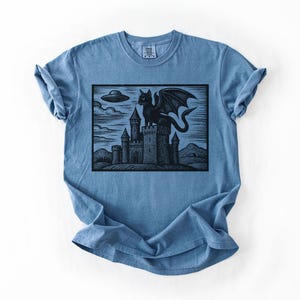May include: Blue t-shirt featuring a black graphic of a winged cat perched atop a castle, with a UFO in the sky. The design is a woodcut style illustration. The shirt has short sleeves and a crew neck.