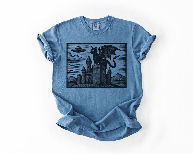 May include: Blue t-shirt featuring a black graphic of a winged cat perched atop a castle, with a UFO in the sky. The design is a woodcut style illustration. The shirt has short sleeves and a crew neck.