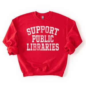 May include: A red sweatshirt with the words "SUPPORT PUBLIC LIBRARIES" in white, distressed, block letters. The sweatshirt has long sleeves and a crew neck. The image is on a white background.