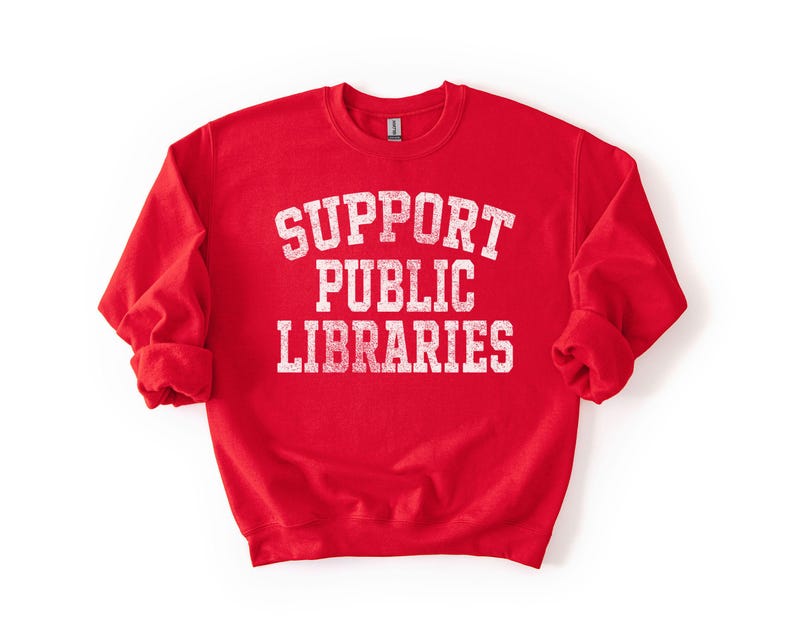 May include: A red sweatshirt with the words "SUPPORT PUBLIC LIBRARIES" in white, distressed, block letters. The sweatshirt has long sleeves and a crew neck. The image is on a white background.