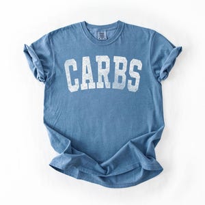 May include: A blue t-shirt with the word "CARBS" printed in large, white, distressed letters. The shirt has short sleeves and a crew neck. The fabric appears soft and slightly faded.