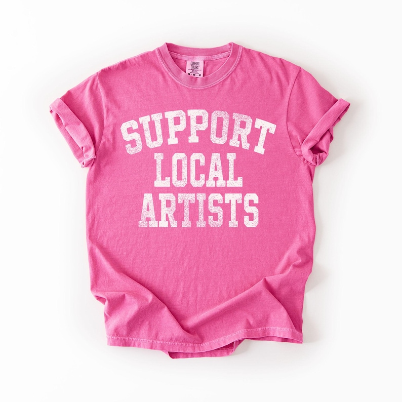 May include: A bright pink t-shirt with the text "SUPPORT LOCAL ARTISTS" in white block letters. The shirt has short sleeves and a crew neck. The fabric appears soft and slightly faded.