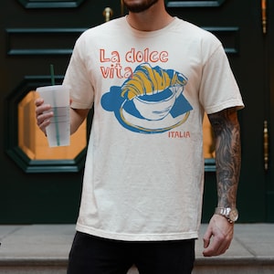 May include: A white t-shirt with a graphic of a croissant in a coffee cup. The text on the shirt reads "La dolce vita" and "ITALIA".