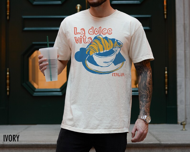 May include: A white t-shirt with a graphic of a croissant in a coffee cup. The text on the shirt reads "La dolce vita" and "ITALIA".
