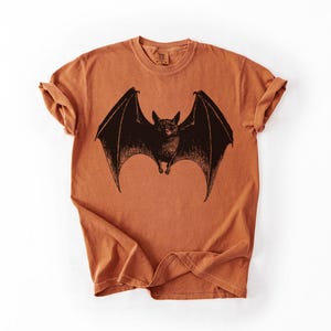 May include: An orange-brown t-shirt featuring a detailed black illustration of a bat with outstretched wings. The bat is centered on the shirt, with the wings spanning across the chest. The shirt has a classic crew neck and short sleeves.