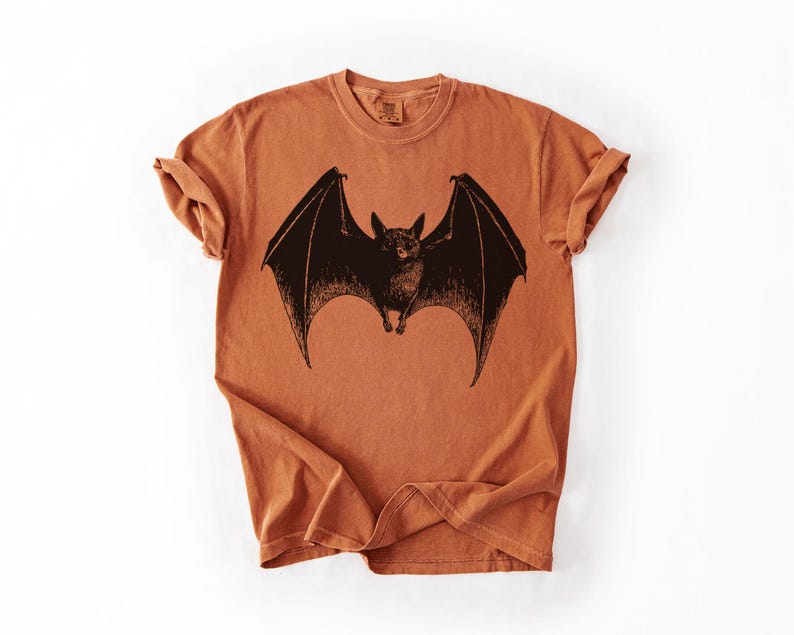 May include: An orange-brown t-shirt featuring a detailed black illustration of a bat with outstretched wings. The bat is centered on the shirt, with the wings spanning across the chest. The shirt has a classic crew neck and short sleeves.