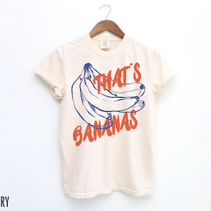 Puede incluir: Camiseta color marfil con un gr&aacute;fico de pl&aacute;tanos azules y rojos y el texto "THAT'S BANANAS". La camiseta est&aacute; colgada en una percha de madera sobre un fondo blanco.