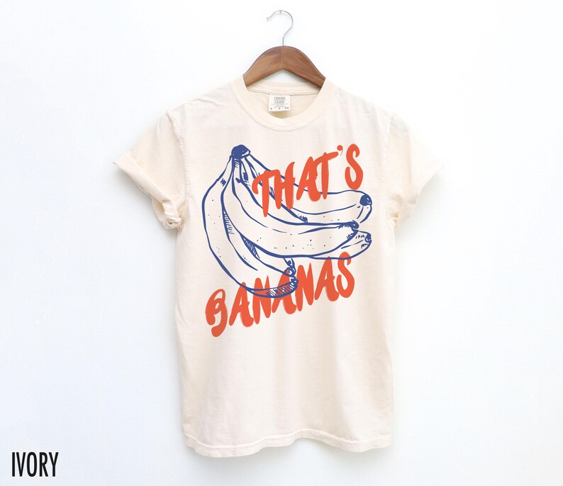 Puede incluir: Camiseta color marfil con un gr&aacute;fico de pl&aacute;tanos azules y rojos y el texto "THAT'S BANANAS". La camiseta est&aacute; colgada en una percha de madera sobre un fondo blanco.