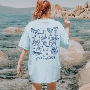 May include: A light blue t-shirt with a graphic design of beach-themed items, including sunglasses, flip-flops, cocktails, and a beach umbrella. The text "Miami Beach Girl's Trip 2025" is printed on the back of the shirt.