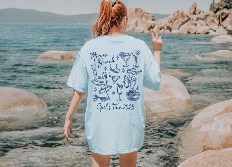 May include: A light blue t-shirt with a graphic design of beach-themed items, including sunglasses, flip-flops, cocktails, and a beach umbrella. The text "Miami Beach Girl's Trip 2025" is printed on the back of the shirt.