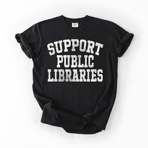 May include: Black t-shirt with the text "SUPPORT PUBLIC LIBRARIES" in white, distressed-style lettering. The shirt is a crew neck and has rolled-up sleeves. The shirt is on a white background.