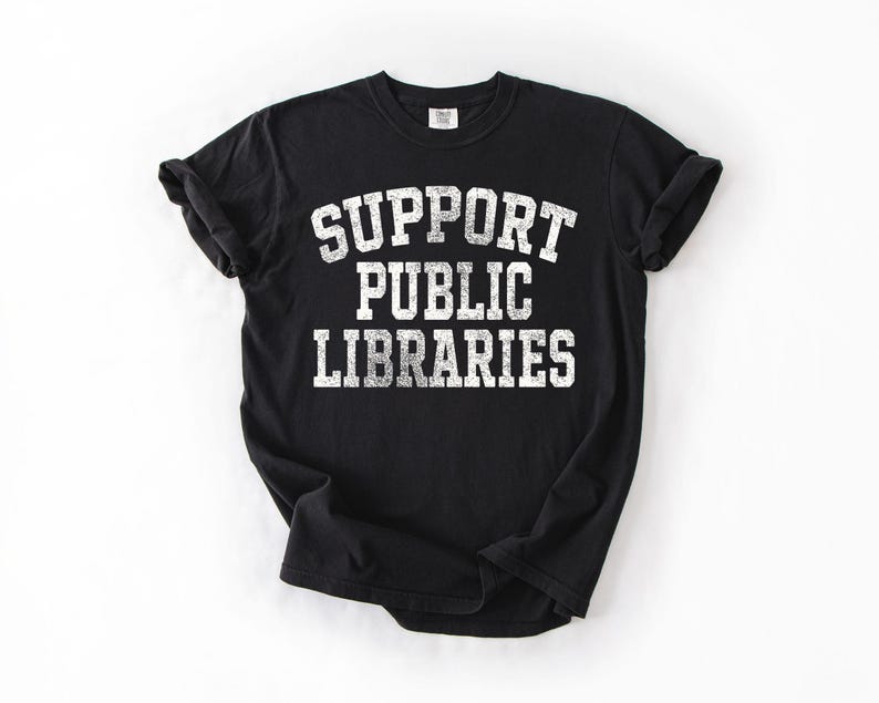 May include: Black t-shirt with the text "SUPPORT PUBLIC LIBRARIES" in white, distressed-style lettering. The shirt is a crew neck and has rolled-up sleeves. The shirt is on a white background.