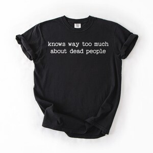Puede incluir: Camiseta negra con el texto blanco "knows way too much about dead people". La camiseta tiene mangas cortas y cuello redondo. La tela parece suave y c&oacute;moda.