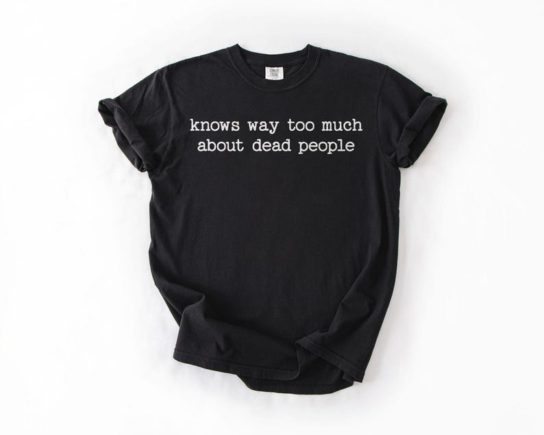 Puede incluir: Camiseta negra con el texto blanco "knows way too much about dead people". La camiseta tiene mangas cortas y cuello redondo. La tela parece suave y c&oacute;moda.