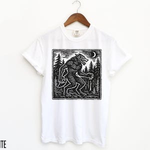 Puede incluir: Camiseta blanca con una ilustraci&oacute;n en blanco y negro al estilo xilograf&iacute;a de un hombre lobo en un bosque bajo una luna creciente. La palabra "WHITE" est&aacute; impresa en la parte inferior. El dise&ntilde;o es de estilo xilograf&iacute;a.