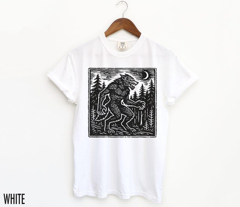 Puede incluir: Camiseta blanca con una ilustraci&oacute;n en blanco y negro al estilo xilograf&iacute;a de un hombre lobo en un bosque bajo una luna creciente. La palabra "WHITE" est&aacute; impresa en la parte inferior. El dise&ntilde;o es de estilo xilograf&iacute;a.