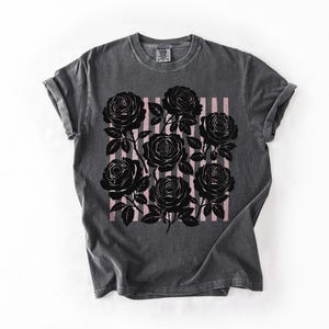 May include: Dark gray t-shirt featuring a black rose graphic with pink vertical stripes. The roses are detailed with leaves and stems. The shirt has a classic crew neck and rolled sleeves.