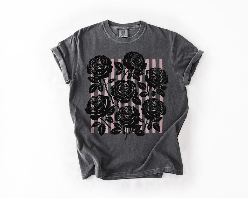 May include: Dark gray t-shirt featuring a black rose graphic with pink vertical stripes. The roses are detailed with leaves and stems. The shirt has a classic crew neck and rolled sleeves.