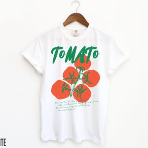May include: White t-shirt with a green and red tomato graphic and the word "TOMATO" in green lettering. The text below the graphic reads "Your tiny tomato. We've got all the inside scoop. The delicious ways to eat the juicy, unsurpassed fruit."