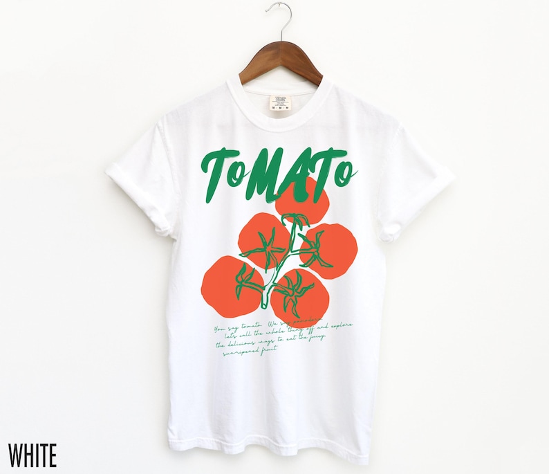 May include: White t-shirt with a green and red tomato graphic and the word "TOMATO" in green lettering. The text below the graphic reads "Your tiny tomato. We've got all the inside scoop. The delicious ways to eat the juicy, unsurpassed fruit."