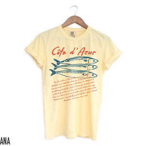 May include: A light yellow t-shirt with the text "C&ocirc;te d'Azur" in red and three blue fish illustrations. Below the text is a paragraph of red text. The shirt is hanging on a wooden hanger.